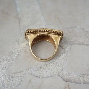 May include: A gold ring with a square-shaped band and a beaded edge. The ring is on a white marble surface.