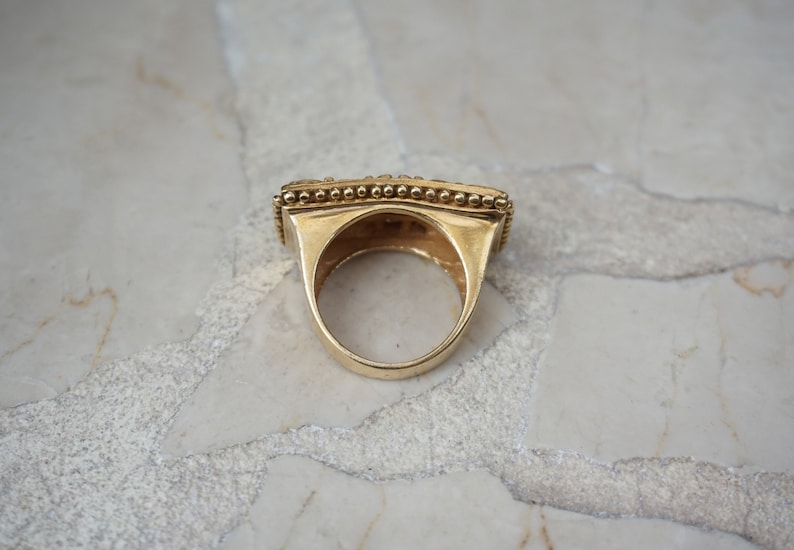 May include: A gold ring with a square-shaped band and a beaded edge. The ring is on a white marble surface.