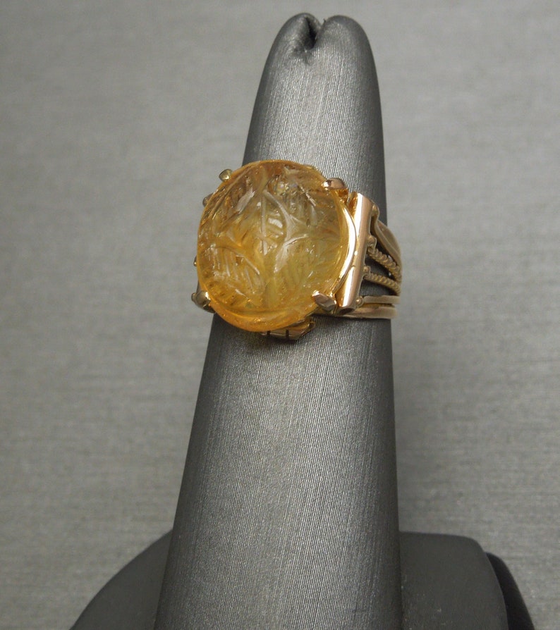 Op de afbeelding: Een gouden ring met een grote, gegraveerde, amberkleurige edelsteen. De ring heeft een gedetailleerd, gevlochten band.
