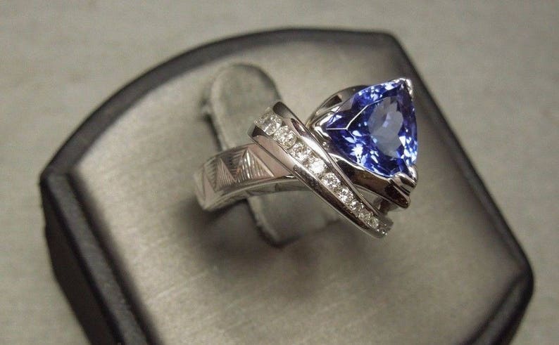 May include: A silver ring with a large, blue, triangular-shaped gemstone surrounded by smaller, white gemstones.