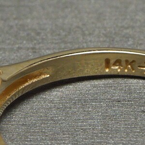 May include: Close-up of a gold ring with a textured design and a band marked "14K". The ring has a series of horizontal ridges on the front. The ring is set against a gray background.
