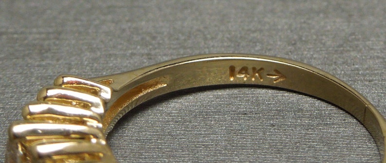 May include: Close-up of a gold ring with a textured design and a band marked "14K". The ring has a series of horizontal ridges on the front. The ring is set against a gray background.