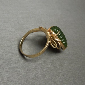 以下が含まれることがあります： 繊細なデザインにセットされた緑色の翡翠の石がついた金の指輪。石には花柄が彫刻されており、指輪には小さなダイヤモンドがセットされています。