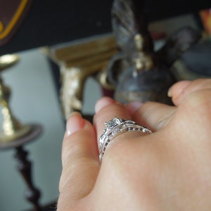 May include: A close-up of a hand wearing a silver engagement ring with a single, clear diamond. The ring has a delicate, intricate design.