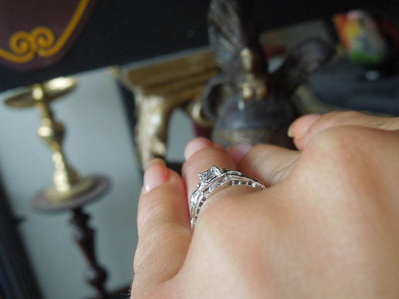 May include: A close-up of a hand wearing a silver engagement ring with a single, clear diamond. The ring has a delicate, intricate design.