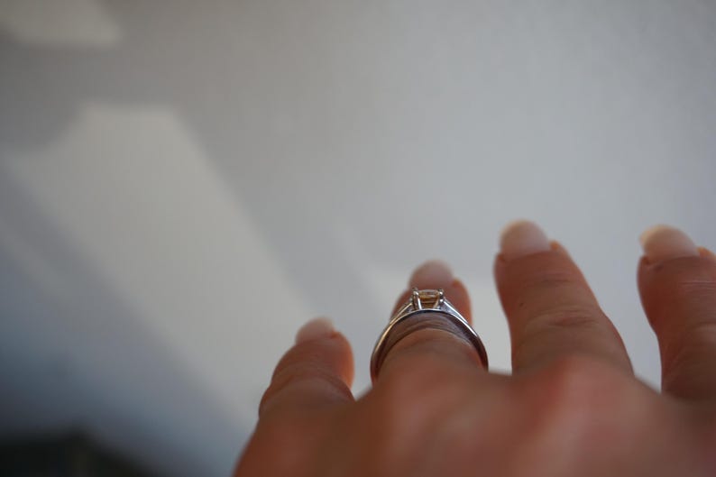 Pode incluir: Um close-up de uma m&atilde;o usando um anel de prata com uma gema transparente. O anel est&aacute; no dedo m&eacute;dio da m&atilde;o.