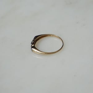 May include: A gold ring featuring a line of small, dark gemstones. The ring is presented on a white surface, highlighting its understated elegance. The band of the ring has a smooth, polished appearance.