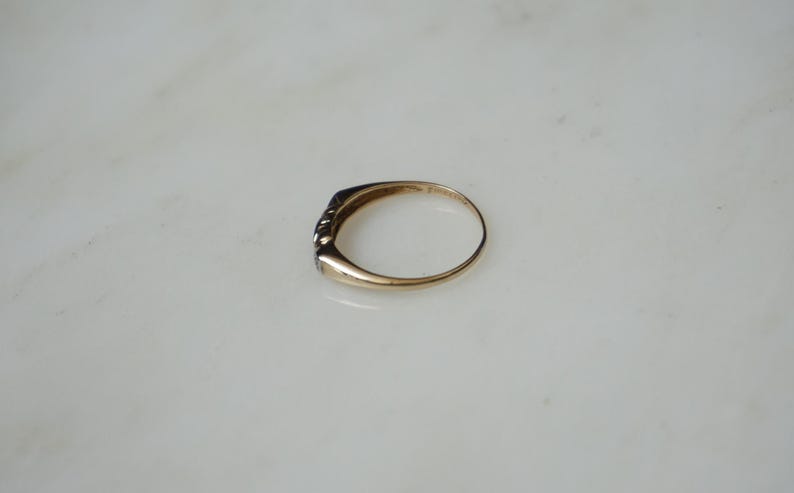 May include: A gold ring featuring a line of small, dark gemstones. The ring is presented on a white surface, highlighting its understated elegance. The band of the ring has a smooth, polished appearance.