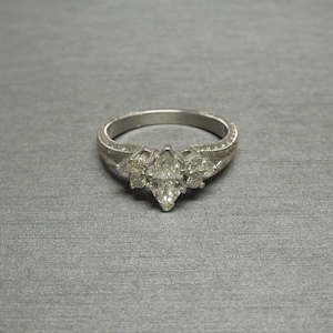 Pode incluir: Um anel de noivado de prata com um diamante central em forma de marquise e dois diamantes menores de cada lado. A banda &eacute; decorada com um delicado design floral.