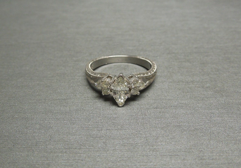 Pode incluir: Um anel de noivado de prata com um diamante central em forma de marquise e dois diamantes menores de cada lado. A banda &eacute; decorada com um delicado design floral.