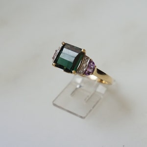 Pu&ograve; includere: Un anello d'oro con una grande pietra preziosa verde smeraldo circondata da diamanti pi&ugrave; piccoli e due pietre di ametista.