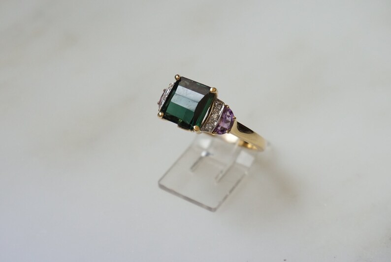 Pu&ograve; includere: Un anello d'oro con una grande pietra preziosa verde smeraldo circondata da diamanti pi&ugrave; piccoli e due pietre di ametista.