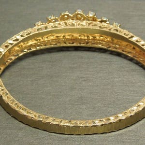 May include: Gold bangle bracelet with a decorative filigree design and a row of clear stones around the top.