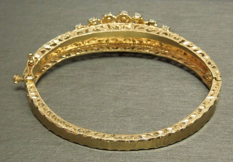 May include: Gold bangle bracelet with a decorative filigree design and a row of clear stones around the top.