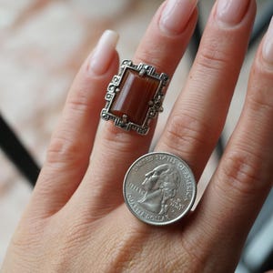 May include: A silver ring with a brown rectangular stone set in a filigree design. The ring is on a person's finger next to a US quarter.