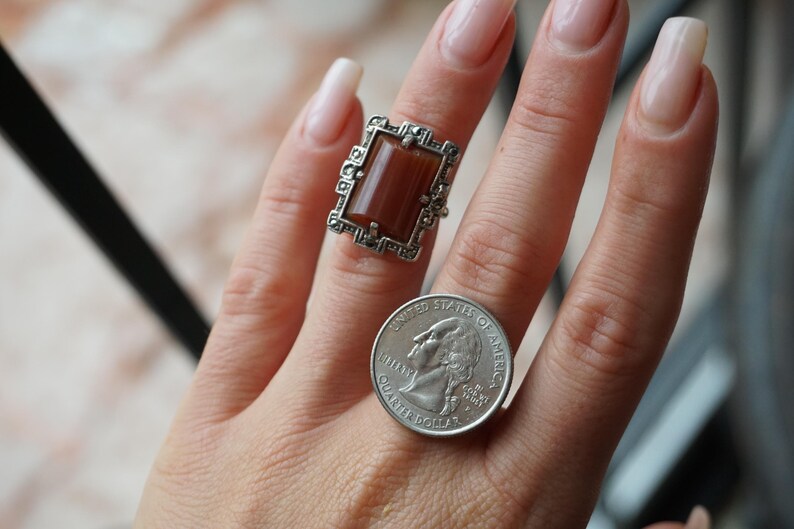 May include: A silver ring with a brown rectangular stone set in a filigree design. The ring is on a person's finger next to a US quarter.