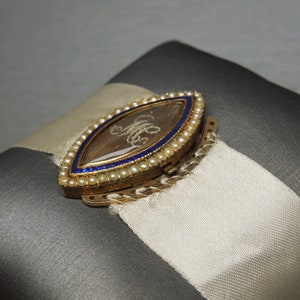 May include: A gold locket with a blue and white enamel border and a small, clear glass window. The locket is decorated with tiny pearls and has a hinged clasp.