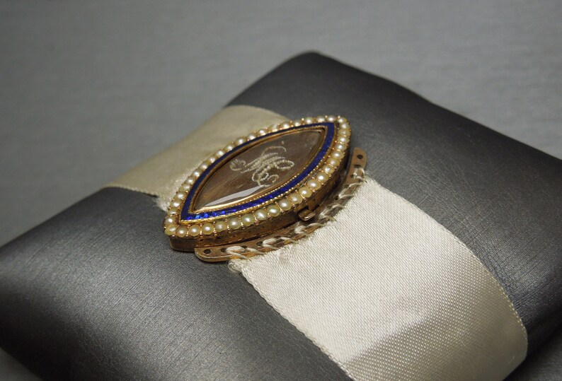 May include: A gold locket with a blue and white enamel border and a small, clear glass window. The locket is decorated with tiny pearls and has a hinged clasp.