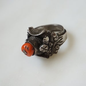 May include: A silver ring with an intricate design featuring a red coral stone. The ring has a wide band and a detailed floral pattern.
