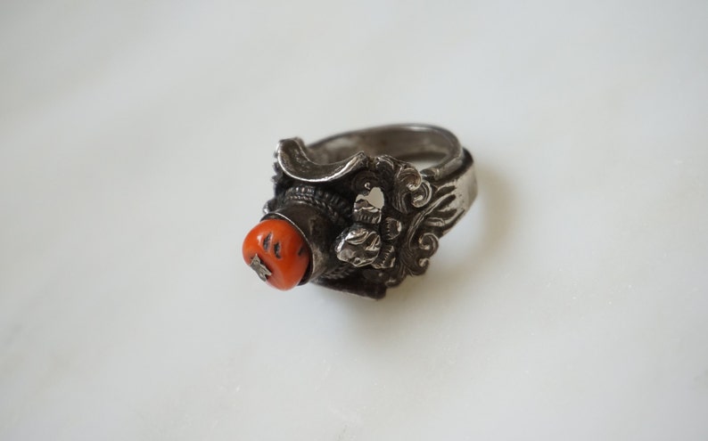 May include: A silver ring with an intricate design featuring a red coral stone. The ring has a wide band and a detailed floral pattern.