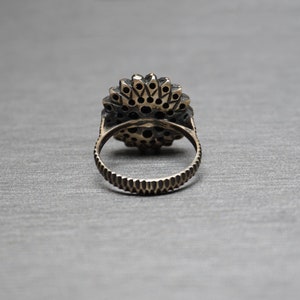 Pu&ograve; includere: Un anello d'argento con una grande pietra preziosa nera rotonda circondata da pietre preziose nere pi&ugrave; piccole. L'anello &egrave; posto su uno sfondo grigio.