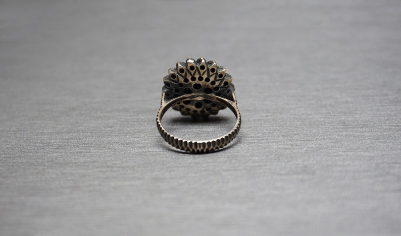 Pu&ograve; includere: Un anello d'argento con una grande pietra preziosa nera rotonda circondata da pietre preziose nere pi&ugrave; piccole. L'anello &egrave; posto su uno sfondo grigio.