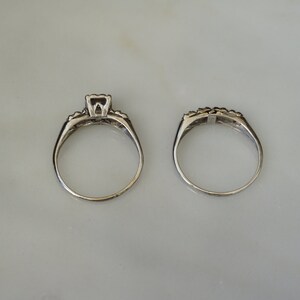 May include: Two silver rings, a wedding set, are displayed on a light-colored surface. The engagement ring features a square-cut center stone and smaller accent stones. The matching band has a row of small stones.