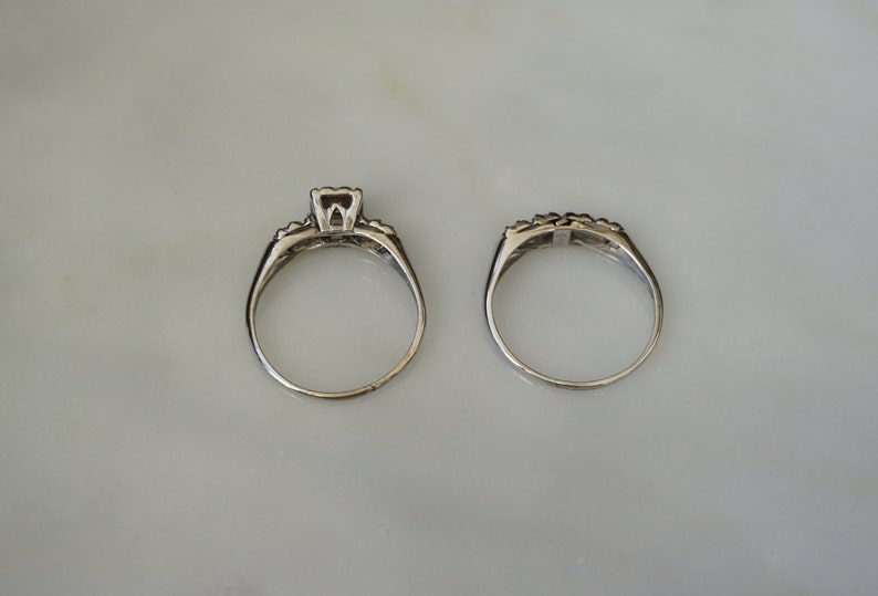 May include: Two silver rings, a wedding set, are displayed on a light-colored surface. The engagement ring features a square-cut center stone and smaller accent stones. The matching band has a row of small stones.