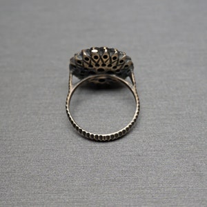 Pu&ograve; includere: Un anello d'argento con una grande pietra preziosa rotonda nera circondata da pietre preziose pi&ugrave; piccole e chiare. L'anello ha una fascia testurizzata.