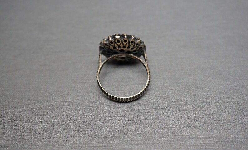 Pu&ograve; includere: Un anello d'argento con una grande pietra preziosa rotonda nera circondata da pietre preziose pi&ugrave; piccole e chiare. L'anello ha una fascia testurizzata.