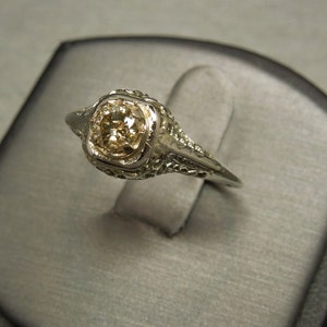 Puede incluir: Un anillo de compromiso vintage con un solo diamante redondo engastado en un dise&ntilde;o de filigrana. El anillo est&aacute; hecho de oro blanco y tiene un dise&ntilde;o delicado e intrincado.
