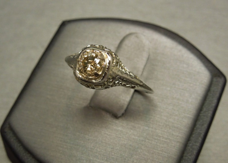 Puede incluir: Un anillo de compromiso vintage con un solo diamante redondo engastado en un dise&ntilde;o de filigrana. El anillo est&aacute; hecho de oro blanco y tiene un dise&ntilde;o delicado e intrincado.