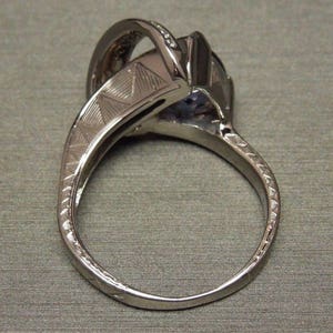 May include: A silver ring with a large purple gemstone and smaller clear gemstones set in a prong setting. The band is textured with a geometric pattern.