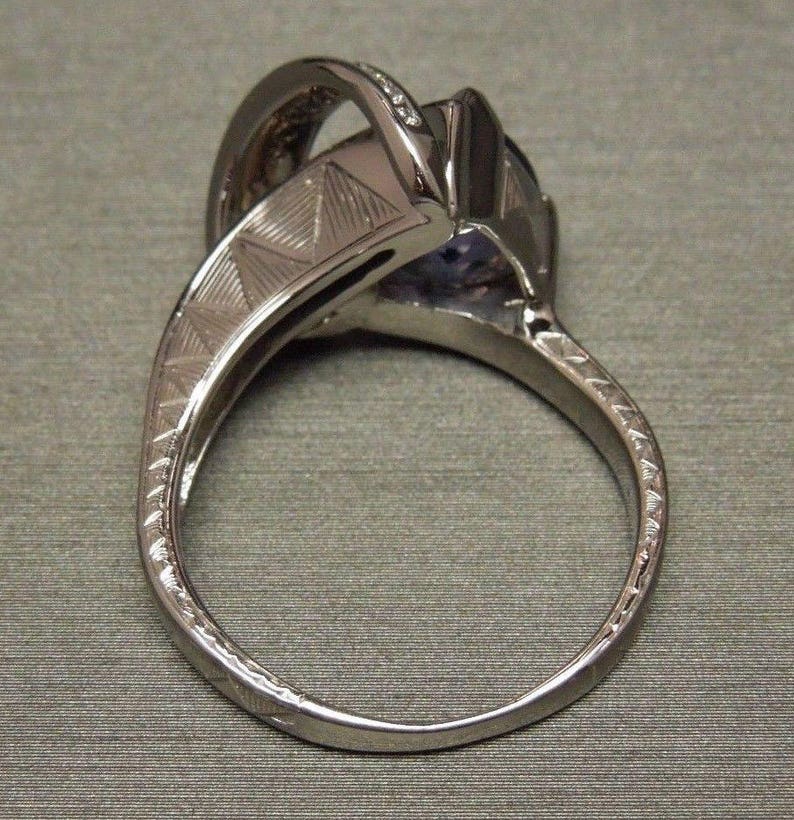 May include: A silver ring with a large purple gemstone and smaller clear gemstones set in a prong setting. The band is textured with a geometric pattern.