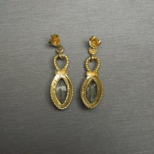 May include: A pair of gold earrings with a teardrop-shaped grey stone set in a gold frame. The earrings have a twisted rope design and a small loop at the top.