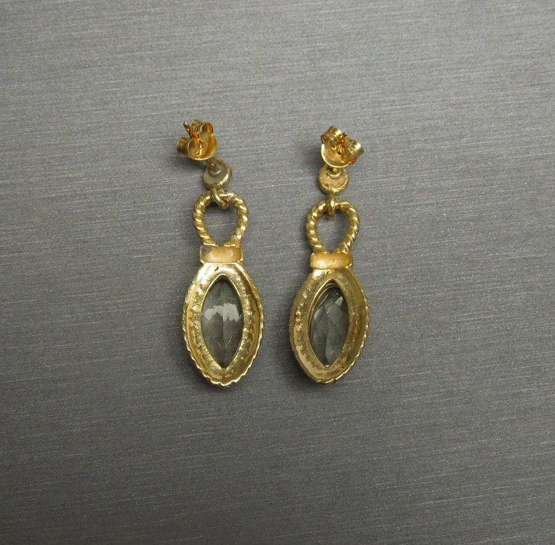 May include: A pair of gold earrings with a teardrop-shaped grey stone set in a gold frame. The earrings have a twisted rope design and a small loop at the top.