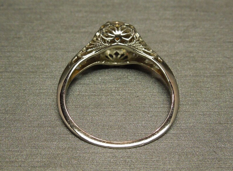 Puede incluir: Un anillo de compromiso de filigrana de plata con una piedra central redonda. El anillo se muestra desde abajo, mostrando los intrincados detalles de la banda.