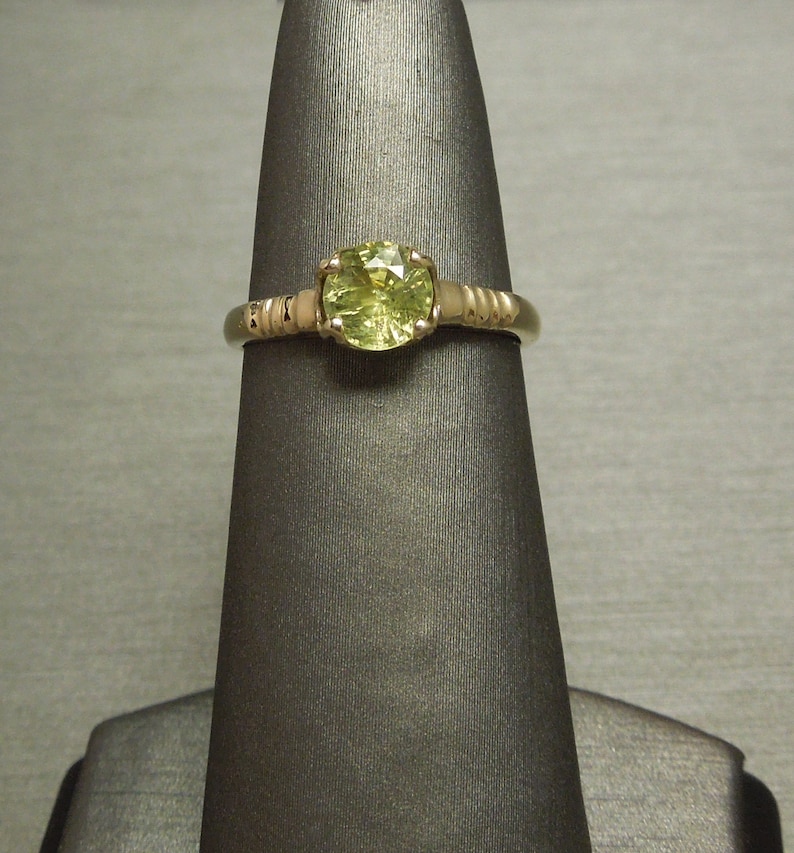 May include: A gold ring with a round, light green gemstone set in a prong setting. The band is textured with a series of small, raised bumps.