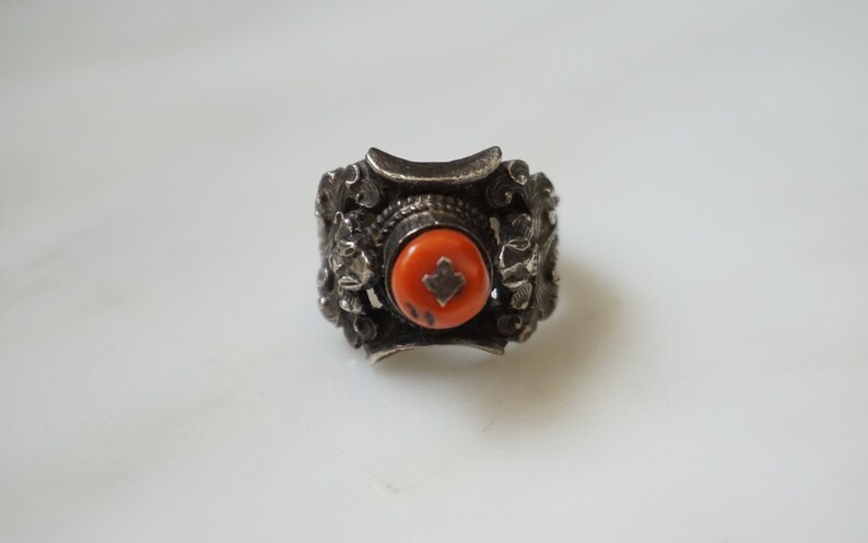 May include: A silver ring with an orange stone set in the center. The ring is decorated with intricate floral designs.