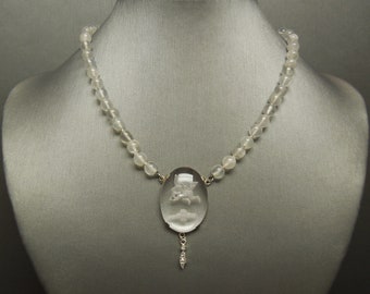 May include: A white glass bead necklace with a carved glass pendant featuring a cherub design. The pendant is attached to a small, clear crystal drop.