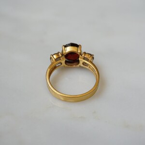 May include: A gold ring with a large red gemstone and two smaller clear gemstones. The ring is on a white surface.