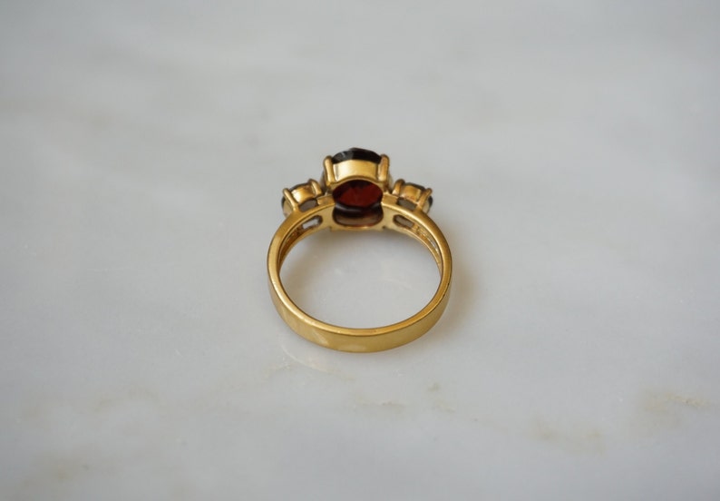 May include: A gold ring with a large red gemstone and two smaller clear gemstones. The ring is on a white surface.