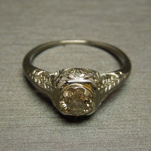 Puede incluir: Un anillo de compromiso vintage con un delicado dise&ntilde;o de filigrana y una piedra central redonda y transparente.