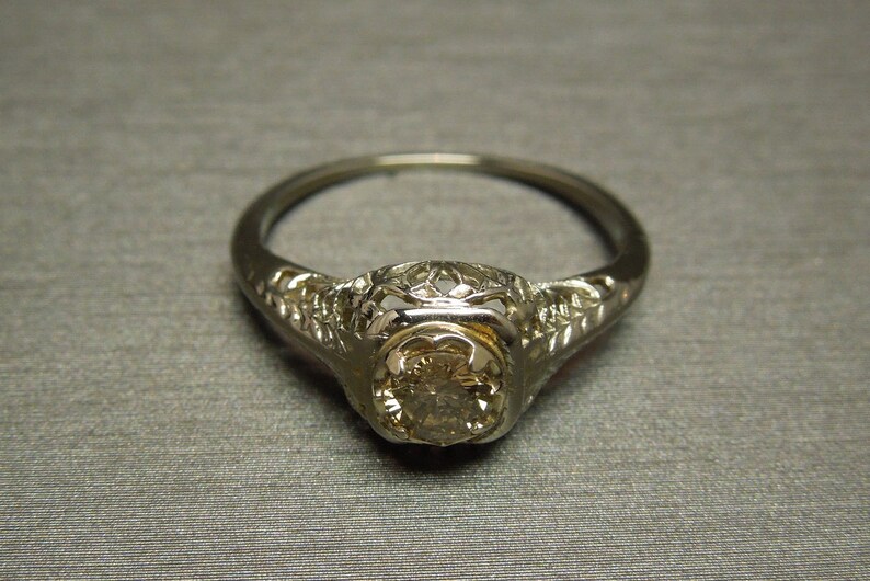 Puede incluir: Un anillo de compromiso vintage con un delicado dise&ntilde;o de filigrana y una piedra central redonda y transparente.