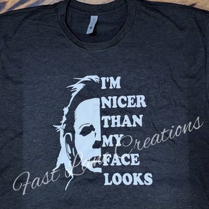 May include: Dark gray t-shirt with a white graphic of a character's face and the text "I'M NICER THAN MY FACE LOOKS." The shirt has a crew neck and short sleeves. The text "Fast Lane Creations" is also printed on the shirt.