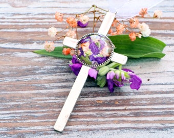 May include: A silver cross pendant with a round glass center filled with pressed purple and white flowers. The pendant is on a wooden surface with white flowers and green leaves.