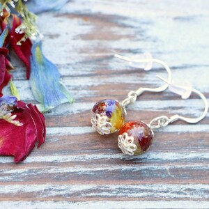May include: A pair of silver earrings with round, colorful resin beads. The beads are filled with pressed flowers and have a silver filigree design around the edge.