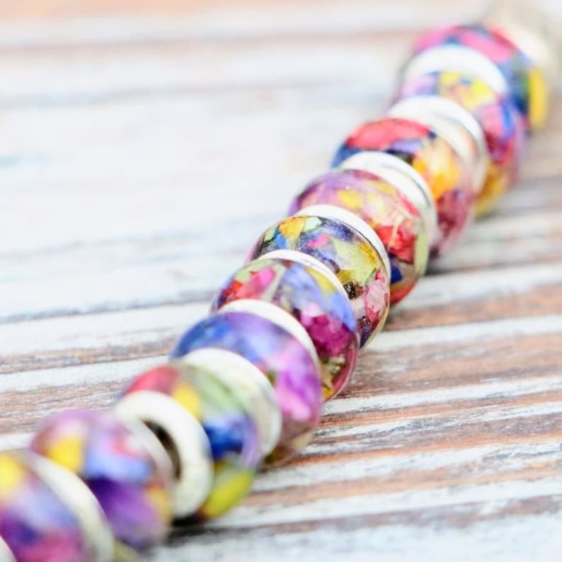 May include: A strand of colorful beads with a floral design. The beads are round, with a clear exterior encasing dried flowers in shades of purple, red, yellow, and blue. Each bead is separated by a silver-colored spacer.