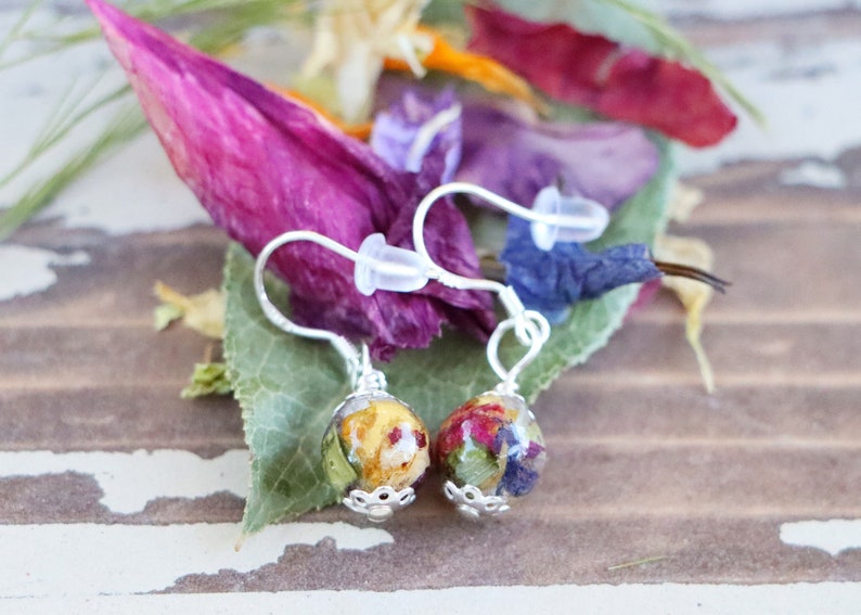 May include: A pair of silver earrings with small round glass beads filled with pressed flowers. The beads are filled with a variety of colors, including yellow, red, and blue.