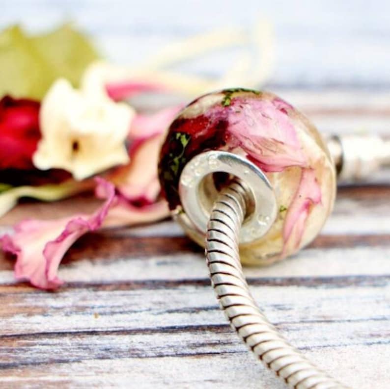 May include: A clear, round bead with dried pink and red flower petals encased inside. The bead is attached to a silver-colored, coiled bracelet. The background features blurred flower petals on a wooden surface.